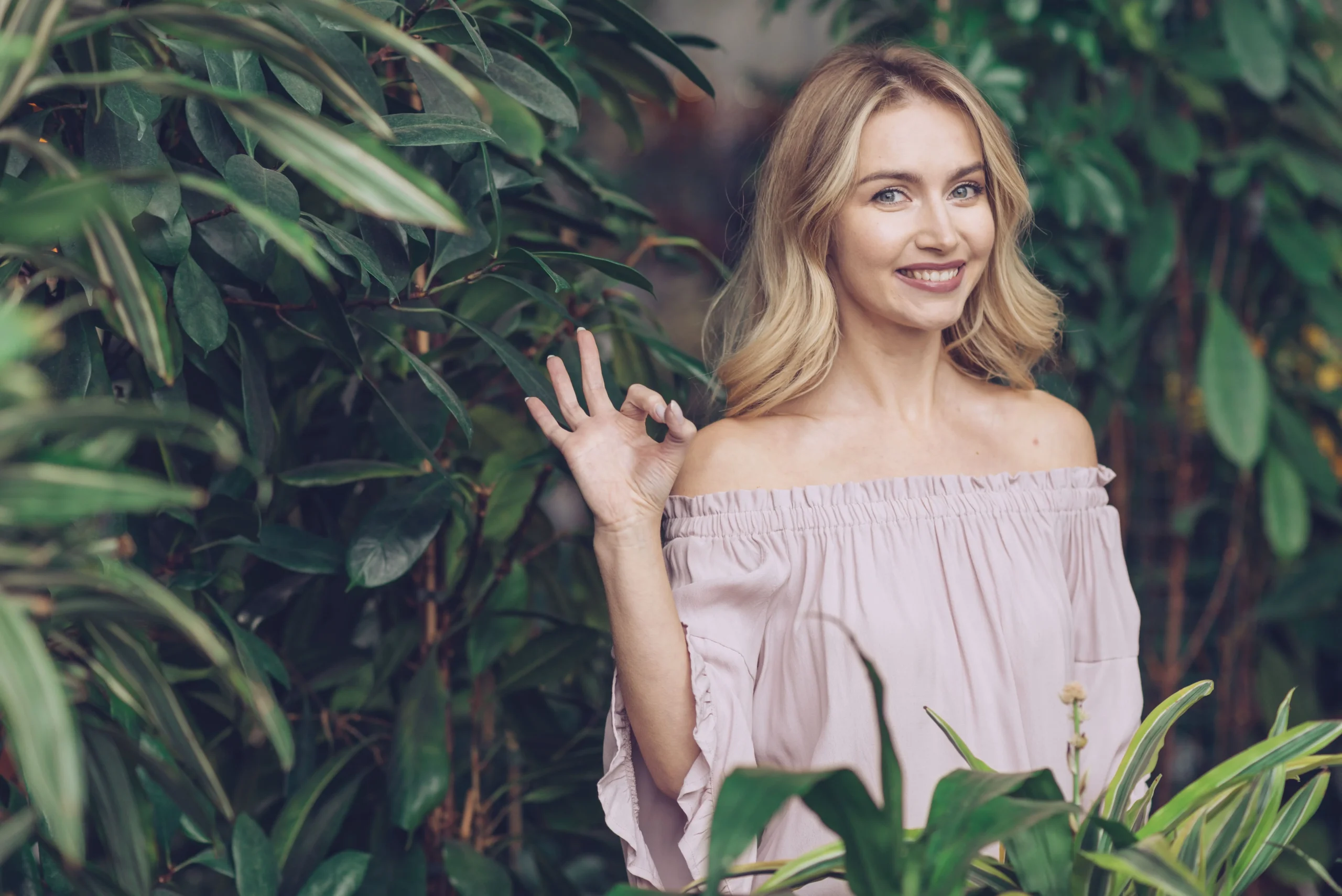 smiling blonde young woman standing front green plant showing ok sign (1)